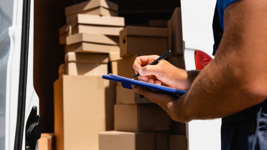 Cropped view of loader writing on clipboard beside cardboard boxes in truck outdoors